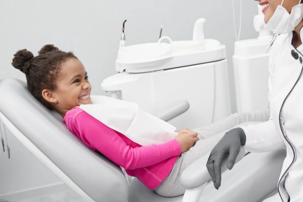 Foto de criança negra sorrindo deitada na cadeira de dentista e uma dentista, estão em um consultório.